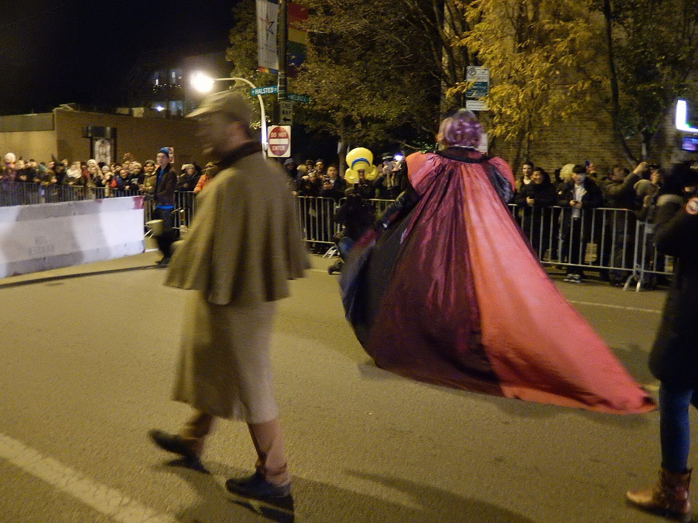 A parade participant in a long red and purple cape walking down a crowded Halsted Street at night
