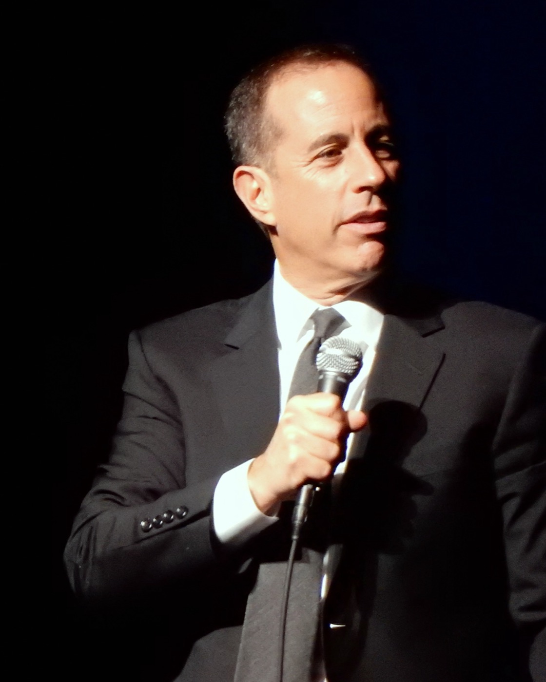 Jerry Seinfeld holding a microphone on stage in a dark suit